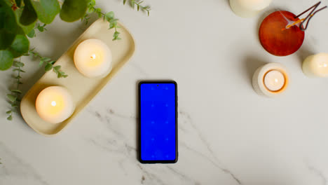 Overhead-View-Looking-Down-On-Still-Life-Of-Blue-Screen-Mobile-Phone-Lit-Candles-And-Incense-Stick-With-Green-Plant-And-Towels-As-Part-Of-Spa-Day-Decor-3